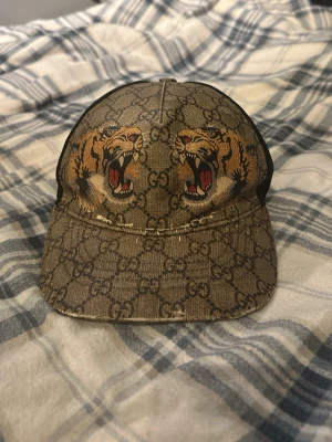 Gucci keps med tigertryck - Snygg keps från Gucci i beige med klassiskt GG-mönster och två stora tigerhuvuden framtill. Kepsen har böjd skärm och justerbar baksida. Tillverkad i Italien och har en exklusiv streetstyle-vibe. Perfekt för dig som vill sticka ut med lyxig accessoar. Tyvärr är loket sönder på boxen.