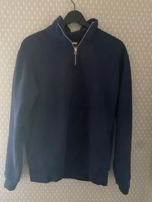 Mörkblå halvzip sweatshirt - Mörkblå sweatshirt med halv dragkedja och hög krage. Tröjan har långa ärmar och ribbade muddar vid ärmslut och nederkant. Perfekt för en avslappnad och stilren look.