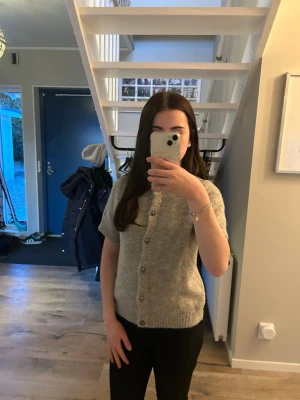 Grå kortärmad kofta från Gina Tricot - Mysig grå kofta med korta ärmar från Gina Tricot. Koftan har rund halsringning, ribbade muddar och knäpps med silverstudds framtill. Perfekt att styla över en t-shirt eller linne för en avslappnad look.