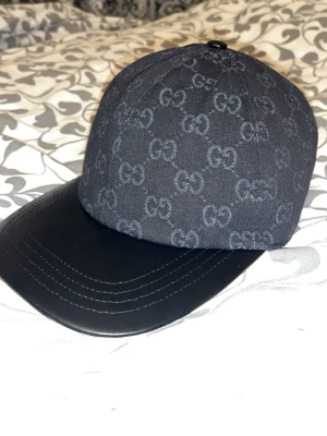 Svart Gucci keps med GG-mönster - Snygg svart keps från Gucci med klassiskt GG-mönster över hela kepsen och en böjd skärm i svart läder. Kepsen har en metallogo på sidan och är tillverkad i tyg med exklusiv känsla. Perfekt för dig som vill ha en stilren och lyxig accessoar. Kan gå ner i pris vid snabb affär 