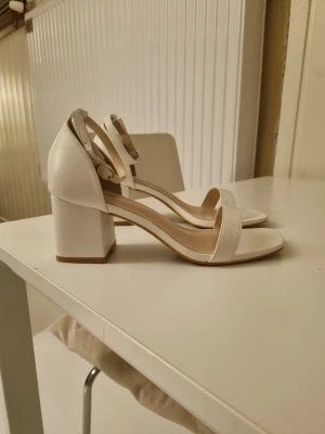 Vita chunky klackpumps med rem - Snygga vita pumps med bred klack och öppen tå. Skorna har en enkel rem över tårna och justerbar ankelrem med spänne. Klacken är chunky och ger stabilitet. Perfekta för dig som vill ha både stil och komfort. Det är storlek 38 men dem är stora i storleken skulle jag säga. Jag brukar ha storlek 38 men dem var förstora på mig så behövde gå ner en storlek. Jag har bara provat dem.