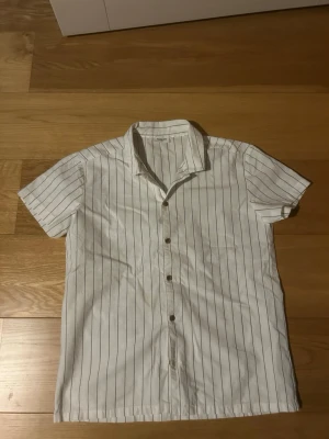 Vit kortärmad randig skjorta - Säljer en vit skjorta med tunna vertikala svarta ränder och korta ärmar. Skjortan har klassisk krage och knäppning framtill med bruna knappar. Perfekt för en avslappnad och stilren look. I storlek 164 och i toppen skick 