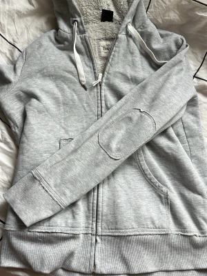 Ljusgrå hoodie från BodyZone - Mysig ljusgrå hoodie med dragkedja från BodyZone. Utrustad med vit fluffig teddyfoder på insidan, huva med dragsko och fickor framtill. Extra detalj med förstärkning på armbågarna. Perfekt för chill dagar och när du vill ha det bekvämt.