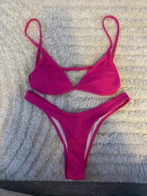 Rosa ribbad bikini med smala band - Trendig rosa bikini med ribbad struktur och smala axelband. Överdelen har triangelformade kupor med uttagbara inlägg och justerbara band. Underdelen är högt skuren för en modern look. 