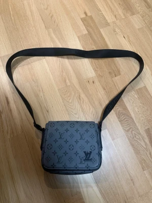 Louis Vuitton axelväska monogram grå - Snygg axelväska från Louis Vuitton i grått med klassiskt monogrammönster. Väskan har en bred, justerbar axelrem och är tillverkad i canvas med detaljer i läder. Perfekt för dig som vill ha en stilren och exklusiv accessoar.