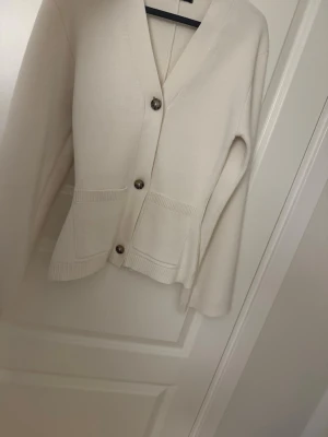 Krämvit kofta från Gina Tricot - Säljer en helt oanvänd snygg krämvit kofta från Gina Tricot med v-ringning och  bruna knappar.