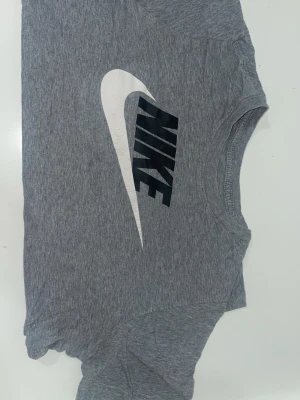 Grå Nike t-shirt med logga - Snygg grå t-shirt från Nike med stor vit och svart logga på bröstet. Klassisk rund halsringning och korta ärmar. Tillverkad i mjuk bomull som känns skön mot huden. Perfekt för en sportig och avslappnad stil.