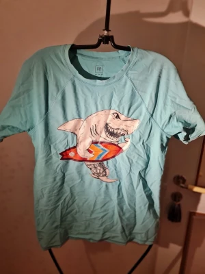 Turkos t-shirt med haj från GAP - Cool turkos t-shirt från GAP med ett stort tryck av en haj som håller en färgglad surfbräda. T-shirten har rund hals och korta ärmar, perfekt för en avslappnad stil. Tillverkad i mjuk bomull som känns skön mot huden.