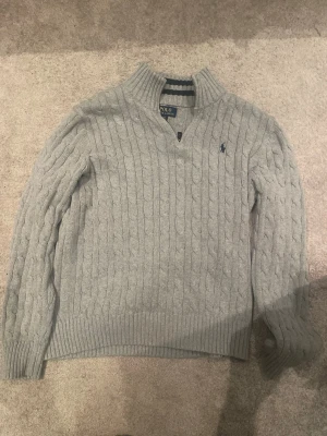 Ljusgrå kabelstickad tröja Polo Ralph Lauren - Ljusgrå kabelstickad tröja från Polo Ralph Lauren med halv dragkedja och broderad logga på bröstet. Tröjan har ribbade muddar och krage, samt klassisk kabelstickad struktur. Perfekt för lager på lager och snygg till jeans.