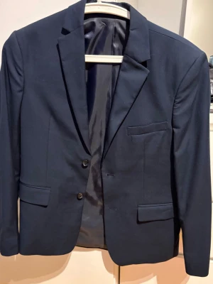 Mörkblå kostym med kavaj och byxor - Snygg mörkblå kostym med klassisk kavaj och matchande byxor. Kavajen har två knappar, tre fickor fram och stilren krage. Byxorna har raka ben och enkel knappstängning.  Passar 164-170. Använd 1 gång vilket syns, backfickorna är inte ens öppnade 