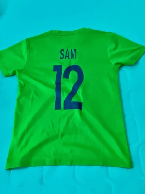 Neongul t-shirt med tryck och nummer - Cool neongul t-shirt med tryck på både fram- och baksida. På ryggen står det 'SAM' och siffran 12 i blått. Framtill finns också siffran 12 och ett vitt emblem. Perfekt för dig som gillar sportig stil.