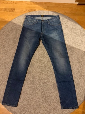 Blå slim fit jeans från Lee - Snygga blå slim fit jeans från Lee med klassisk femficksdesign och diskreta slitningar framtill. Jeansen har normal midja och raka ben, tillverkade i ett mjukt denimtyg som ger en bekväm känsla. Perfekta för en avslappnad och trendig look.