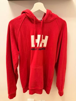 Röd hoodie från Helly Hansen - Röd hoodie från Helly Hansen med stor vit HH-logga och svart text på bröstet. Klassisk känguruficka framtill och justerbar huva med dragsko. Perfekt för en avslappnad och sportig stil.