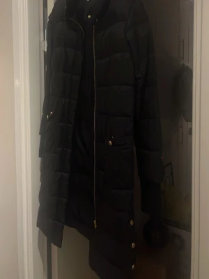 Svart lång dunjacka från Busnel - Snygg svart lång dunjacka från Busnel med guldfärgade knappar och dragkedja. Jackan har quiltad design, fickor med lock och tryckknappar samt en patch med logga på ärmen. 