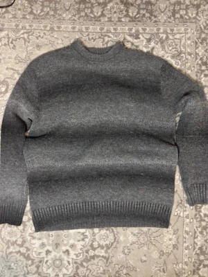 Mörkgrå stickad tröja 157 - Mysig mörkgrå stickad tröja från 157 med rund halsringning och ribbade muddar vid ärmslut och nederkant. Perfekt för kyliga dagar och enkel att matcha med olika outfits. Klassisk och stilren design utan mönster.