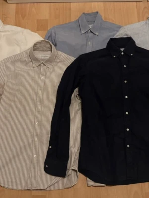 John Henric skjortor, strl S - Säljer ett set med fem skjortor i olika färger och mönster. Skjortorna har klassisk krage och knäppning framtill. Färgerna är vit, ljusblå, svart, beige med ränder och ljusgrå med ränder. Perfekta för en stilren och enkel look.