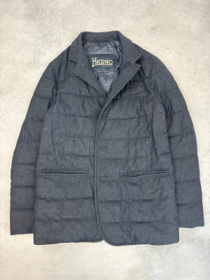 Mörkgrå quiltad dunjacka från Herno - Stilren mörkgrå dunjacka från Herno med quiltad design och klassisk krage. Jackan har två framfickor och en bröstficka, samt diskreta sömmar för en clean look. Perfekt för kalla dagar när du vill vara både varm och snygg.