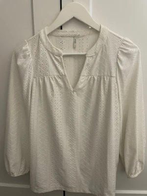 Vit blus med broderade detaljer från ONLY - Snygg vit blus från ONLY med broderade hålmönster och v-ringning. Blusen har puffiga långärmar och en avslappnad passform. Tillverkad i ett lätt och luftigt material som ger en romantisk vibe.