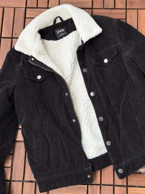 Svart manchesterjacka från ZARA - Säljer en svart manchesterjacka från ZARA med vit fuskpälsfoder och krage. Jackan har tryckknappar, två bröstfickor med lock och två sidofickor. Perfekt för dig som vill ha en stilren och varm cardiganjacka med retrovibbar. Är gjord för män, så kan upplevas som lite större, men jag har även haft den som kvinna. Riktigt varm, sambon har haft hela vintern!