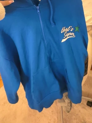 Blå North West hoodie med tryck - Blå hoodie med dragkedja och huva från North West. På bröstet står det 'that's Super' med en grön stjärna och på ryggen finns ut ett stort vitt tryck med texten 'North West'. Hoodien har känguruficka och är gjord i mjukt material.