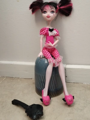 Monster High docka Draculaura - Säljer en Monster High docka av karaktären Draculaura. Hon har svart och rosa hår i tofsar, rosa prickig pyjamas med spets och svarta detaljer samt rosa tofflor med kattansikten. En svart borste i form av en kista ingår.