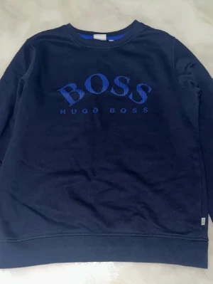 Mörkblå sweatshirt från Hugo Boss - Snygg mörkblå sweatshirt från Hugo Boss med stor BOSS-logga i blått framtill. Tröjan har rund halsringning, långa ärmar och är tillverkad i mjukt material. Perfekt för en avslappnad och stilren look.