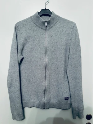 Grå stickad ziptröja - Snygg grå stickad tröja från Jack & Jones med hel dragkedja framtill och hög krage. Tröjan har en minimalistisk look och en liten diskret logga vid nederkanten. Perfekt för lager på lager under kyliga dagar. XS men sitter som S