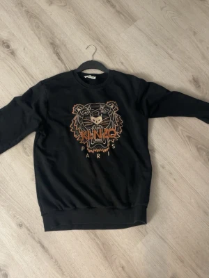 Svart Kenzo sweatshirt med tigertryck - Svart sweatshirt från Kenzo med ikoniskt tigerhuvud broderat framtill och texten 'Kenzo Paris'. Tröjan har rund halsringning, långa ärmar och ribbade muddar vid ärmslut och nederkant. Perfekt för en trendig och avslappnad stil. Det står Storlek XXL men är prkt L