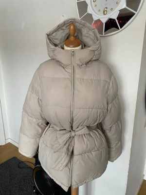 Beige pufferjacka med knytskärp - Snygg beige pufferjacka med huva och knytskärp i midjan. Jackan har en rak, puffig passform och stängs med dragkedja framtill. Perfekt för kalla dagar när du vill hålla dig varm och samtidigt se trendig ut.