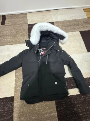 Svart parkas från Moose Knuckles - Säljer en svart parkas från Moose Knuckles med stor huva och vit fuskpäls. Jackan har flera fickor med tryckknappar och den ikoniska metall-loggan på ärmen. Perfekt för kalla dagar och riktigt snygg streetstil.