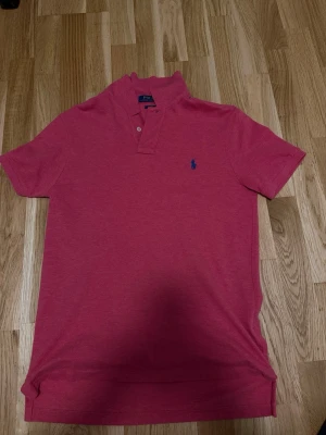 Rosa pikétröja från Ralph Lauren - Klassisk rosa pikétröja från Ralph Lauren med korta ärmar, krage och två knappar framtill. Tröjan har en blå broderad logga på bröstet och är gjord i mjuk bomull som känns skön mot huden.