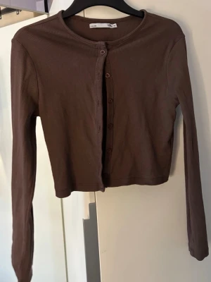 Brun ribbad cropped cardigan - Säljer en brun ribbad cardigan med lång ärm och rund hals. Modellen har knappar framtill. 