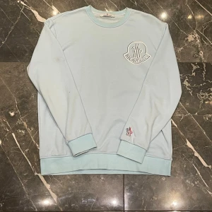 Ljusblå sweatshirt från Moncler - Snygg ljusblå sweatshirt från Moncler med stort broderat Moncler-märke på bröstet och en mindre logga på ärmen. Tröjan har rund halsringning, ribbade muddar och avslappnad passform. Perfekt för en chill och trendig look. Tröjan är smutsig och har fläckar på vissa ställen som på kragen och 2 fläckar på ärmen. 