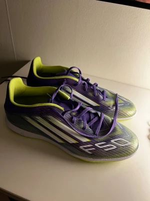 Adidas F50 lila och gula fotbollsskor - Säljer ett par Adidas F50 fotbollsskor.        ‼️Endast andvända en futsalcup så bra skick ‼️ Skorna har snörning, låg profil och en sula anpassad för inomhusspel. Ovandelen är i syntetmaterial med grafiskt mönster och klassiska Adidas-ränder på sidan. Perfekta för dig som vill ha snabba fötter på planen.