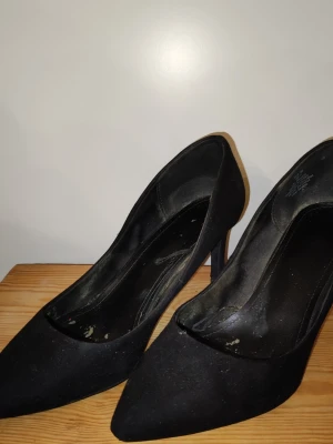 Svarta klassiska pumps med spetsig tå - Svarta pumps med spetsig tå och hög klack. Skorna har en stilren och tidlös design med slät yta och innersula i svart. Perfekta för dig som gillar en elegant och klassisk look. Materialet ser ut att vara syntet eller textil.