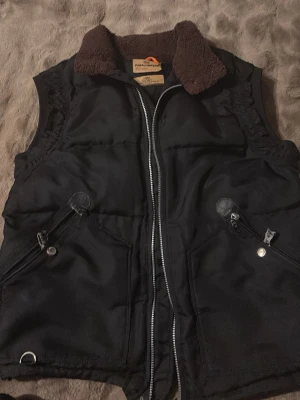 Svart dunväst från Parajumpers - Stilren svart dunväst från Parajumpers Masterpiece Series Anthology med mjuk brun fuskpäls på kragen, två snedställda fickor med dragkedja och tryckknapp samt robust dragkedja framtill. Perfekt för lager-på-lager och streetwear. A kopia 