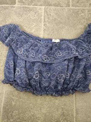 Blå offshoulder spetstopp - Söt blå offshoulder topp med volang och blommig spets. Croppad modell med resår i nederkant och ärmar för en bekväm passform. Perfekt för dig som vill ha en romantisk och trendig look.