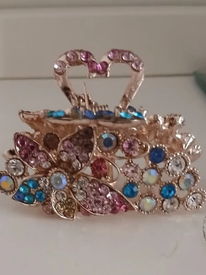 Hårklämma med fjäril och strass - Säljer en glittrig hårklämma i guldfärgad metall med dekorativa fjärilar och hjärtdetalj. Klämman är täckt av färgglada strasstenar i blått, rosa, vitt och gult som ger ett lyxigt intryck. Perfekt accessoar för att piffa upp frisyren.