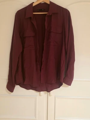 Vinröd skjorta med fickor - Säljer en vinröd skjorta med två bröstfickor och klassisk krage. Skjortan har långa ärmar och knappar framtill. Lite oversize. Aldrig använd