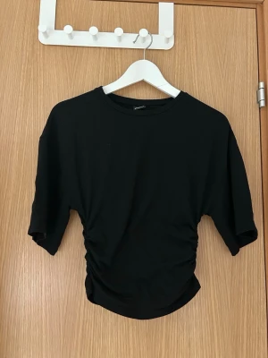 Svart topp med kort ärm - En svart topp från Gina Tricot med korta ärmar och rund hals. Toppen har snygga rynkdetaljer i sidorna som ger en cool och trendig look. Perfekt att styla med jeans eller kjol för en enkel men stilren outfit.