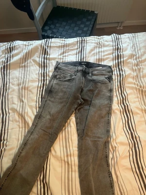 Gråa Replay Anbass jeans - Säljer ett par gråa Replay Anbass jeans i slim fit-modell. Klassisk femficksdesign med dragkedjegylf och snygga slitningar. Perfekta för dig som gillar en modern och stilren look. Materialet är mjukt och stretchigt för extra komfort.