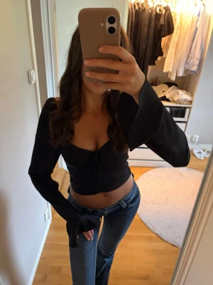Fest topp - Trendig svart croppad topp med långa vida ärmar och snygg knytning framtill. Djup v-ringning och figurnära passform ger en modern look. Perfekt att styla med jeans eller kjol för en cool outfit.