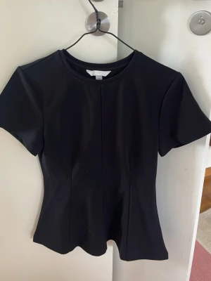 Svart topp från H&M med kort ärm - En stilren svart topp från H&M med rund halsringning och korta ärmar. Toppen har en figurnära passform och är tillverkad i ett mjukt, stretchigt material som känns bekvämt mot huden. Perfekt att matcha med jeans eller kjol för en clean look.