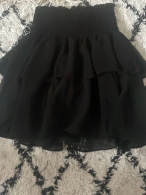 Svart volangkjol från Made by Chelsea - Supersnygg svart kortkjol från Made by Chelsea i silke. Kjolen har bred smock i midjan och flera lager volanger som ger en riktigt fin och fluffig look. Perfekt att styla med en enkel topp eller stickad tröja.