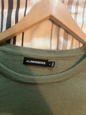 Olivgrön stickad tröja J.Lindeberg - Snygg olivgrön stickad tröja från J.Lindeberg med rund halsringning och diskret logga på bröstet. Tröjan har långa ärmar och ribbade muddar vid ärmslut och nederkant. Perfekt för lager på lager och enkel att matcha.
