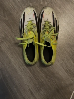 Adidas f50 elite - Säljer ett par Adidas f50 fotbollsskor i vitt och gult med svarta detaljer och snörning. Skorna har klassiska dobbar för spel på gräs och är tillverkade i lätt syntetmaterial för maxad fart på planen. Perfekta för dig som vill sticka ut med både stil och speed.