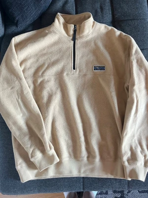 Beige fleece tröja med halv dragkedja - Mysig beige fleece tröja med halv dragkedja och hög krage. Tröjan har ett litet märke på bröstet och ribbade muddar vid ärmslut och nederkant. Perfekt för lager på lager och chill dagar.
