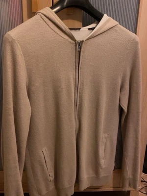 Beige hoodie - Säljer en stilren beige hoodie med dragkedja framtill och två fickor. Hoodien har en klassisk huva och är tillverkad i ett mjukt, stickat material som känns skönt mot huden. Perfekt för dig som gillar en clean och enkel look.