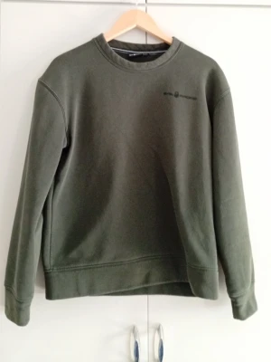 Mörkgrön sweatshirt  - Stilren mörkgrön sweatshirt från sailracing med rund halsringning och diskret logga på bröstet. Tröjan har en avslappnad passform, ribbade muddar vid ärmslut och nederkant samt mjukt material som känns skönt mot huden.