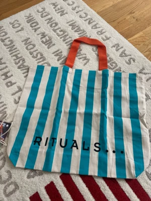 Randig shoppingväska från Rituals - Snygg shoppingväska från Rituals med vertikala turkosa och vita ränder. Väskan har orange handtag och är tillverkad i tyg, perfekt för att bära med sig till skolan eller stan. Texten 'RITUALS...' står tryckt på framsidan. Samarbete med Tiny Miracles.